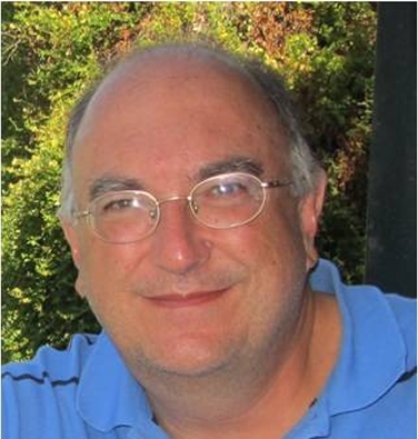 Hombre con gafas y barba, canoso con brazos cruzados y camiseta de médico blanca con boligrafos en el bolsillo en el exterior de un hospital